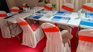A conference room with white tables, blue placemats, and chairs with orange sashes. - Ohm Village Inn (Kitui)