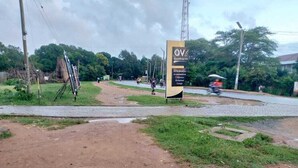 A dirt path leading to a signboard with text, surrounded by greenery and a road with vehicles. - Ohm Village Inn (Kitui)