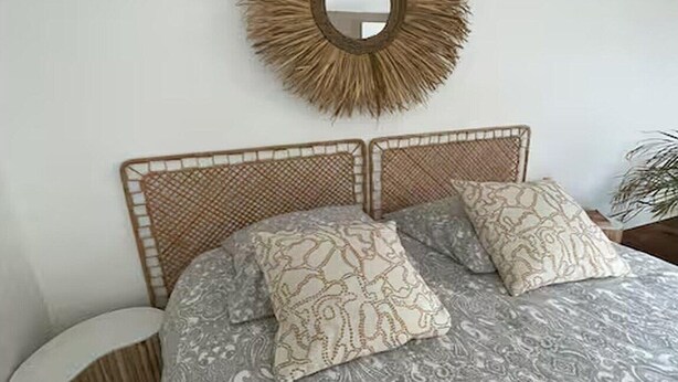 Ein Bett mit einem gemusterten Überwurf, ein runder Spiegel mit strukturierter Rahmen und ein Rattan-Kopfstück.