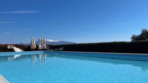 Una piscina con sillones de descanso y sombrillas, un seto y una montaña al fondo.