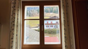 Een kamer met een houten plafond, een raam met uitzicht op een huis en landschap, en een radiator onder het raam.