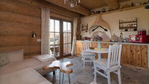 Un intérieur en bois comprenant un canapé, des fauteuils, une table, un lustre et un coin cuisine avec des étagères et une cuisinière.