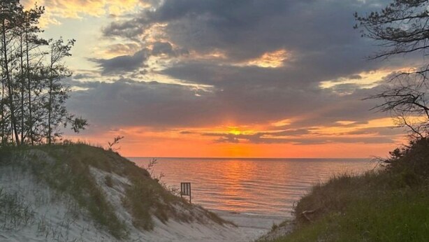 Une plage avec des dunes et une chaise, un coucher de soleil sur l’eau et un ciel nuageux.