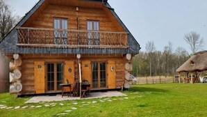 Une cabane en rondins dotée d’un balcon, d’une terrasse en pierre et, au fond, d’une construction au toit de chaume.