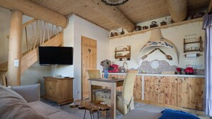 Une pièce chaleureuse avec un escalier en bois, une télévision, une table à manger et un coin cuisine doté de meubles en bois et d’un poêle décoratif.