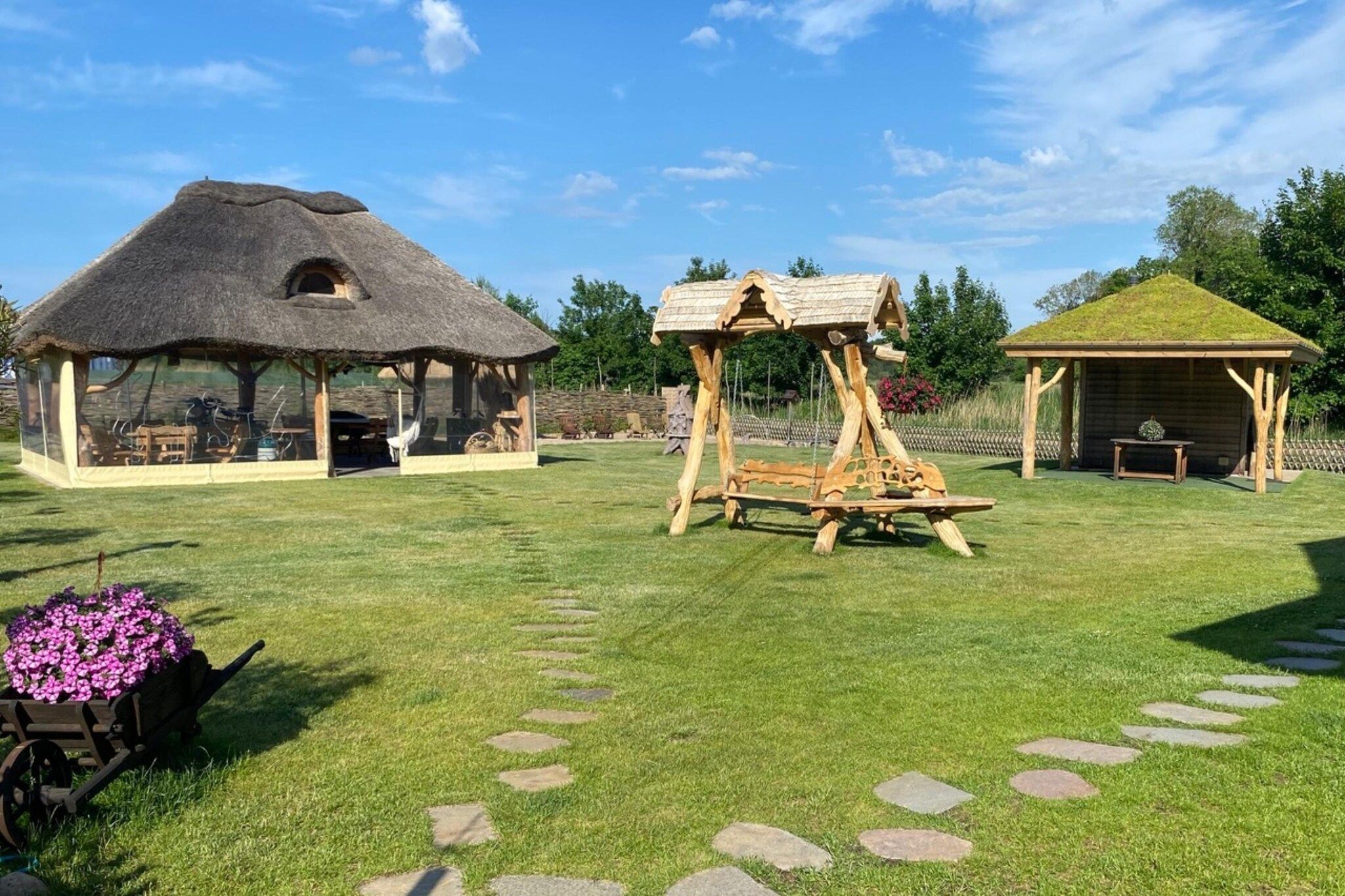 A rustic outdoor setting with a thatched-roof structure, a wooden swing set, and a small wooden building.