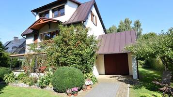 Une maison à deux étages, dotée d’un toit en pente, d’un garage et d’un jardin bien entretenu où poussent diverses plantes et fleurs.