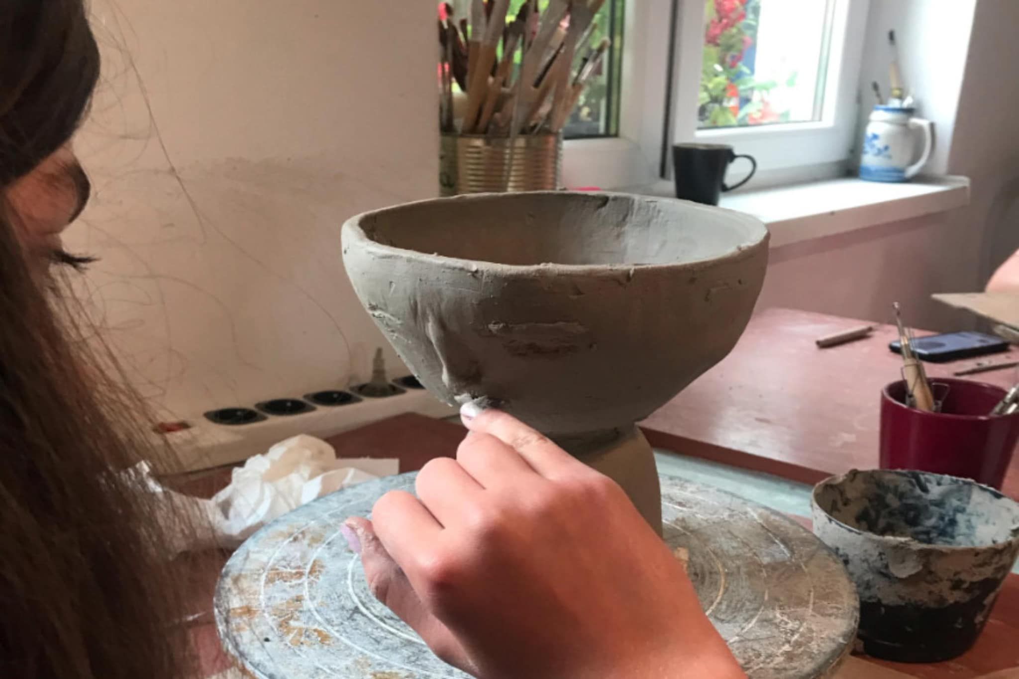 En person arbetar med ett keramikprojekt, och på ett bord framför personen står en skål. Runt omkring finns olika verktyg och material.