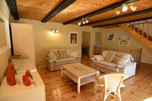 A cozy living room with two sofas, a coffee table, and a wicker chair. There is a staircase with wooden railings and a ceiling with exposed beams.