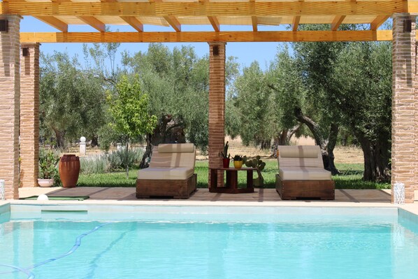 A pool area with two lounge chairs, a brick pillar, and a pergola overhead.