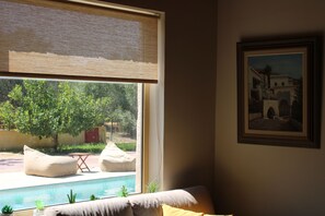 A room with a view of a swimming pool, a sofa, a framed painting, and a window with a blind.