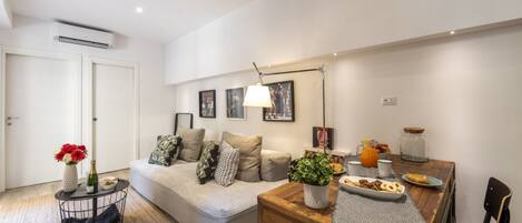 A modern living room with a sofa, a dining table with breakfast items, a coffee machine, and a plant.