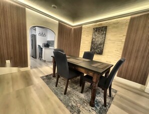 Ein Essbereich mit Holztisch und -stühlen, im Hintergrund eine Küche und eine Wand mit Struktur, an der ein gerahmtes Kunstwerk hängt.