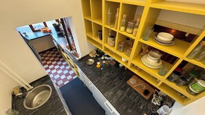 Una cocina con piso a cuadros, un lavamanos y un estante amarillo con frascos y vajilla.