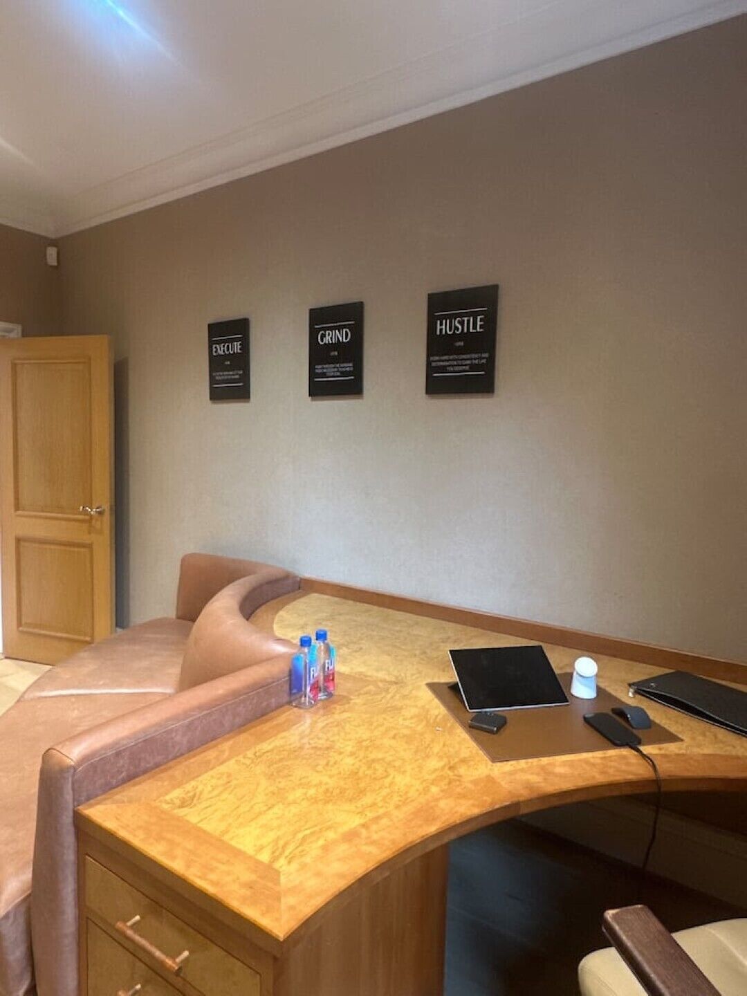A hotel room with a curved wooden desk, a laptop, a tablet, and a smartphone on the desk. There are three motivational signs on the wall.
