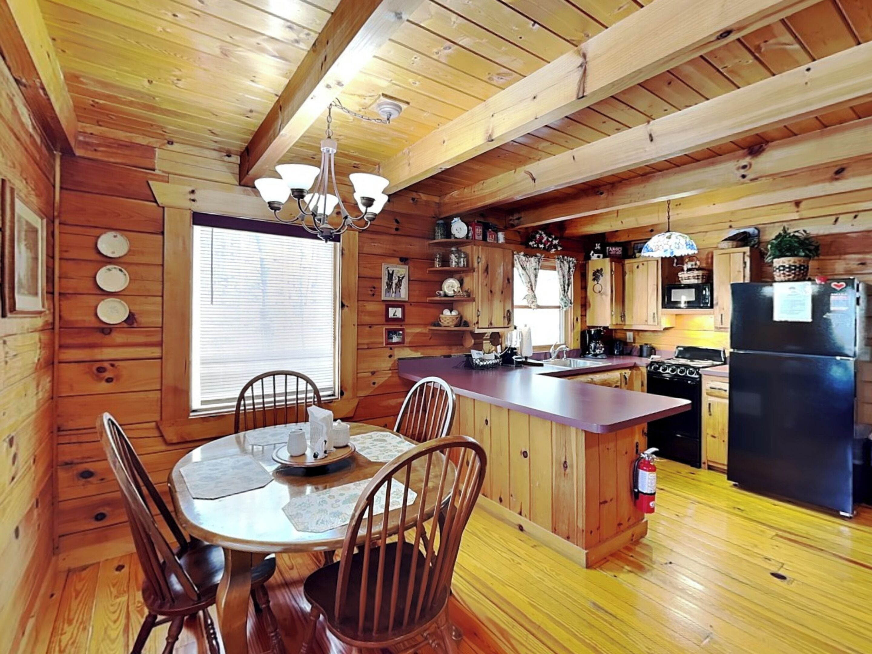 Interno di una baita in legno con tavolo da pranzo, sedie, cucina completa di piano cottura, frigorifero e pensili in legno.