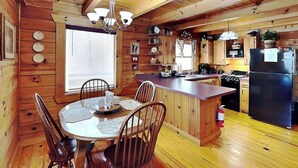 Ein Holzblockhaus-Interieur mit Esstisch, Stühlen, einer Küche inklusive Herd, Kühlschrank und Holzschränken.