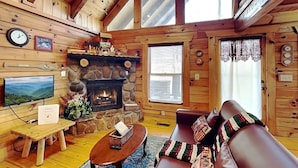 Ein Blockhaus-Interieur mit Kamin aus Stein, Holzwänden, einem Fernseher, einer Couch und einem Esstisch.