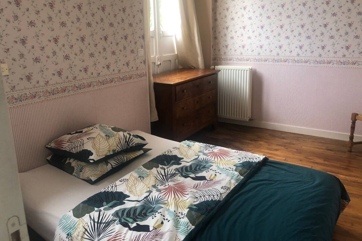 A bedroom with floral wallpaper, a wooden dresser, a radiator, and a bed with a leaf-patterned bedspread.