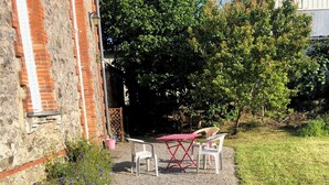 A gravel patio with a red table and two chairs, a stone wall, and a grassy area. - Belle Maison dans une ancienne école (Chemille-en-Anjou)