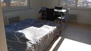 Ein Schlafzimmer mit Bett, Schreibtisch und Stühlen, einem Fernseher und großen Fenstern mit Blick auf eine Stadt und Berge.