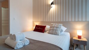 Ein ordentlich bezogenes Bett, gerollte Handtücher, ein rotes Kissen mit Aufschrift, ein flauschiges Kissen, eine Nachttischlampe und eine gestreifte Wand.