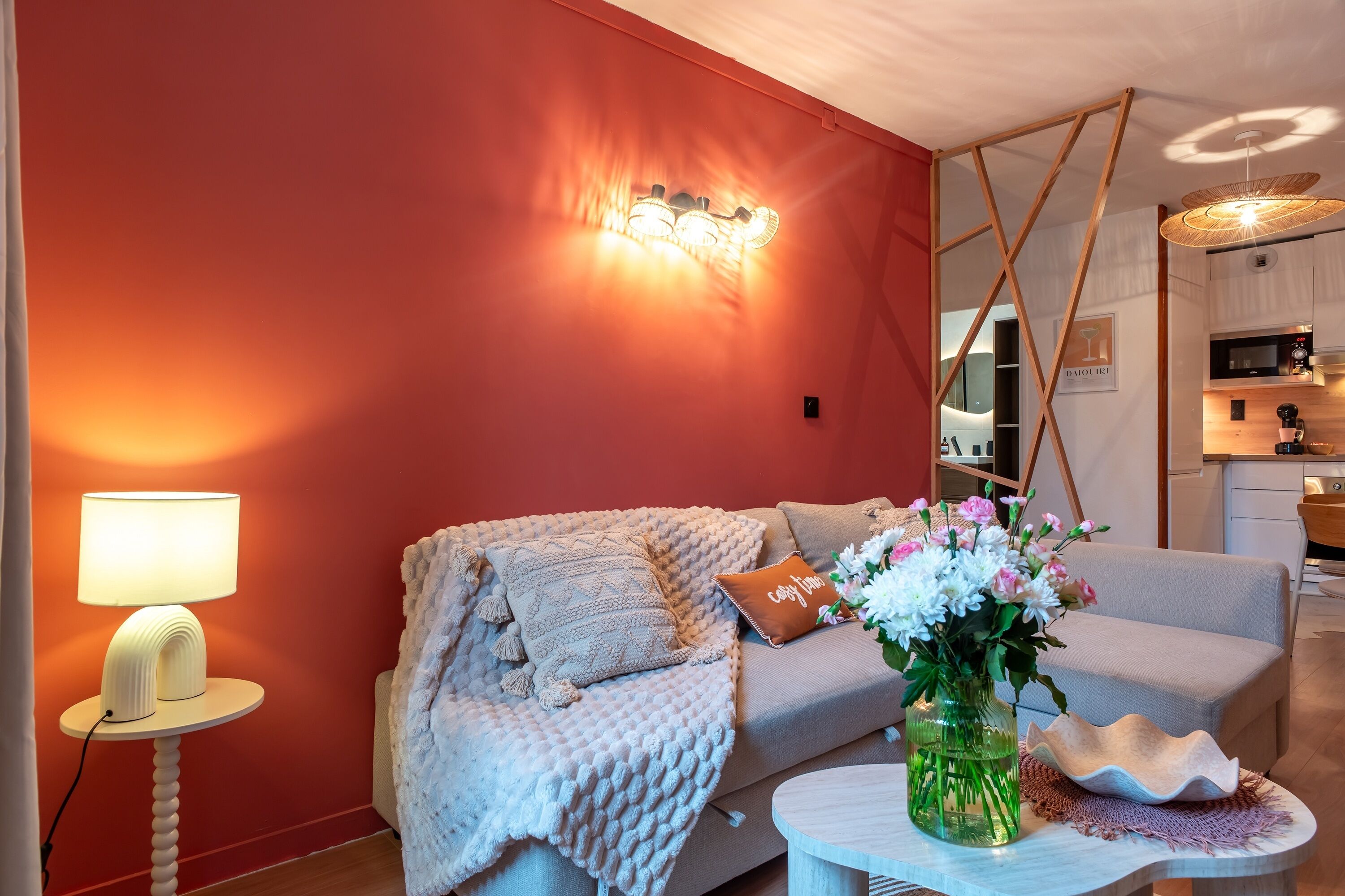 Een moderne woonkamer met een rode muur, een bank, een lamp, een vaas met bloemen en een keuken in de achtergrond.