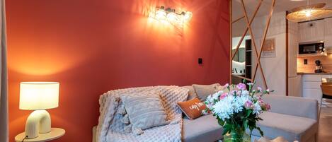 Een moderne woonkamer met een rode muur, een bank, een lamp, een vaas met bloemen en een keuken in de achtergrond.