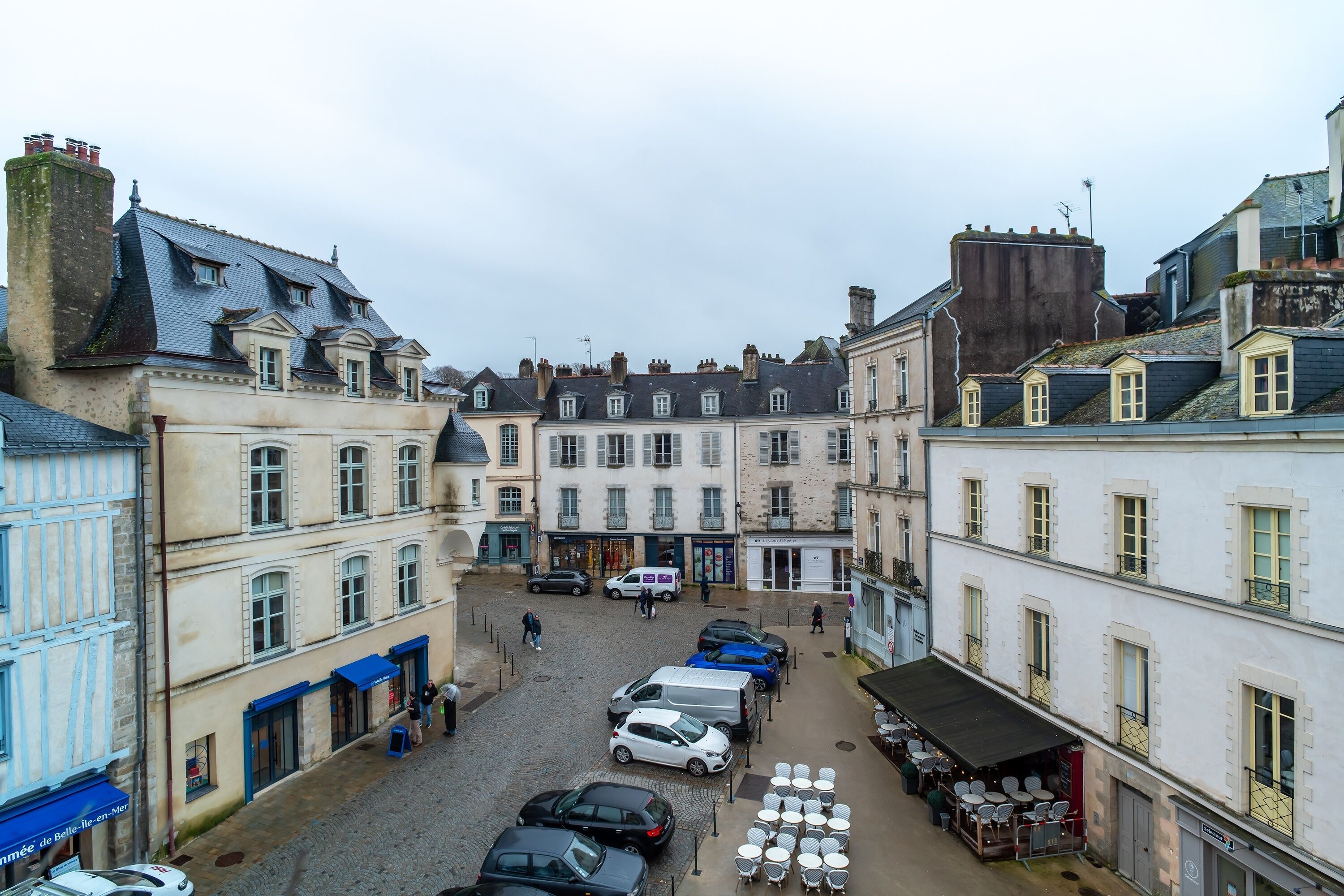A cobblestone street with buildings, parked cars, and outdoor seating.