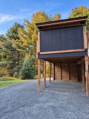 Un bâtiment en bois, doté d’un toit sombre et soutenu par des piliers en bois, est construit sur une surface de gravier, avec des arbres en arrière-plan.