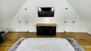 A room with a sloped ceiling, a white wall with a mounted television, a wooden bench, and a patterned rug. - Private Barn Loft w/ King Bed | Cozy Forest Escape (Parry Sound)
