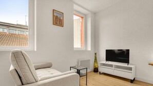 Uma sala de estar moderna com um sofá, uma TV em um suporte branco, uma mesinha e uma janela com vista para edifícios.