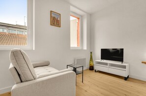 A modern living room with a sofa, a TV on a white stand, a small table, and a window with a view of buildings. - Piccolo - Studio calme et lumineux au cœur de la ville (Toulouse)