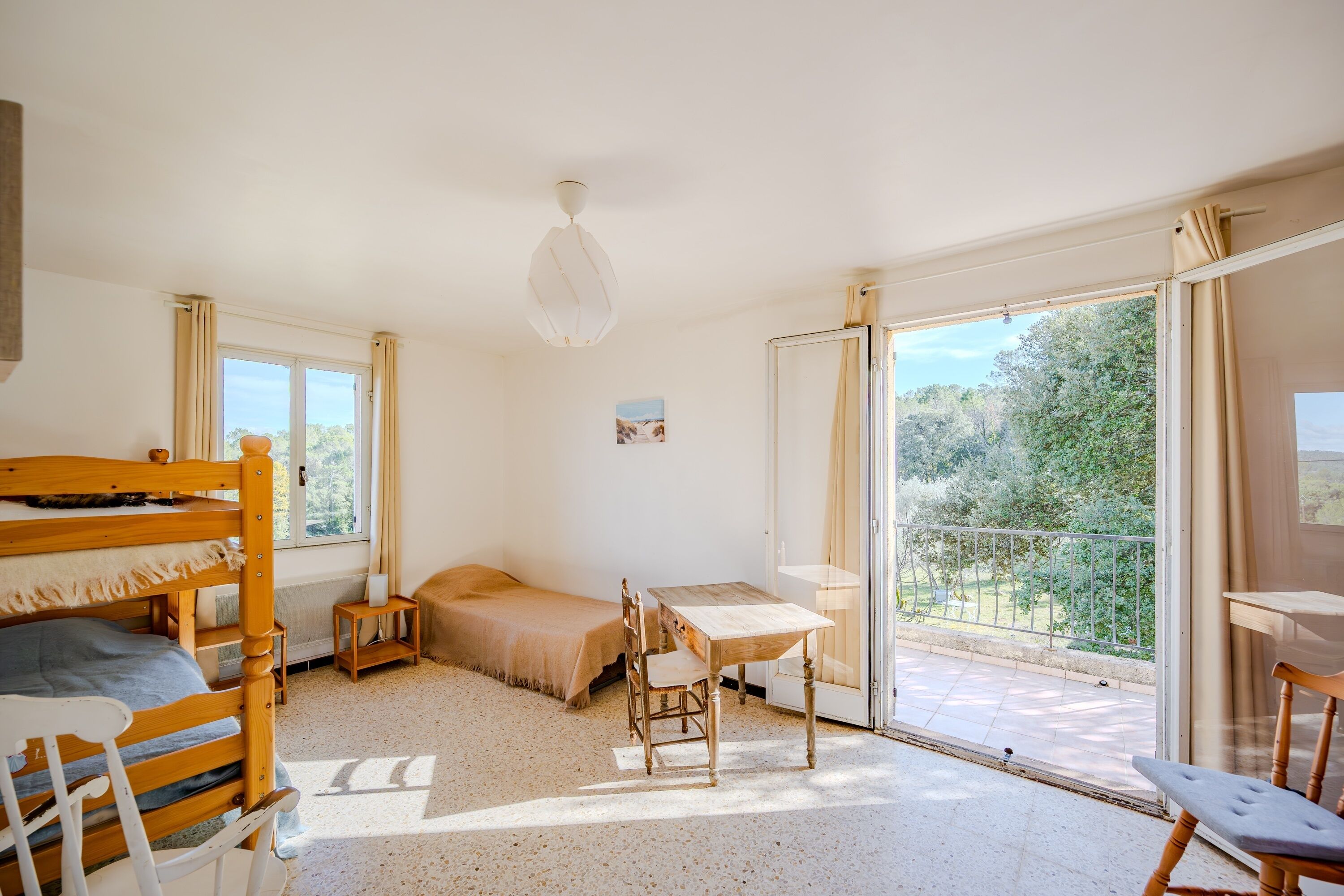 A room with a bunk bed, a small table, a chair, and a balcony with a view of greenery.