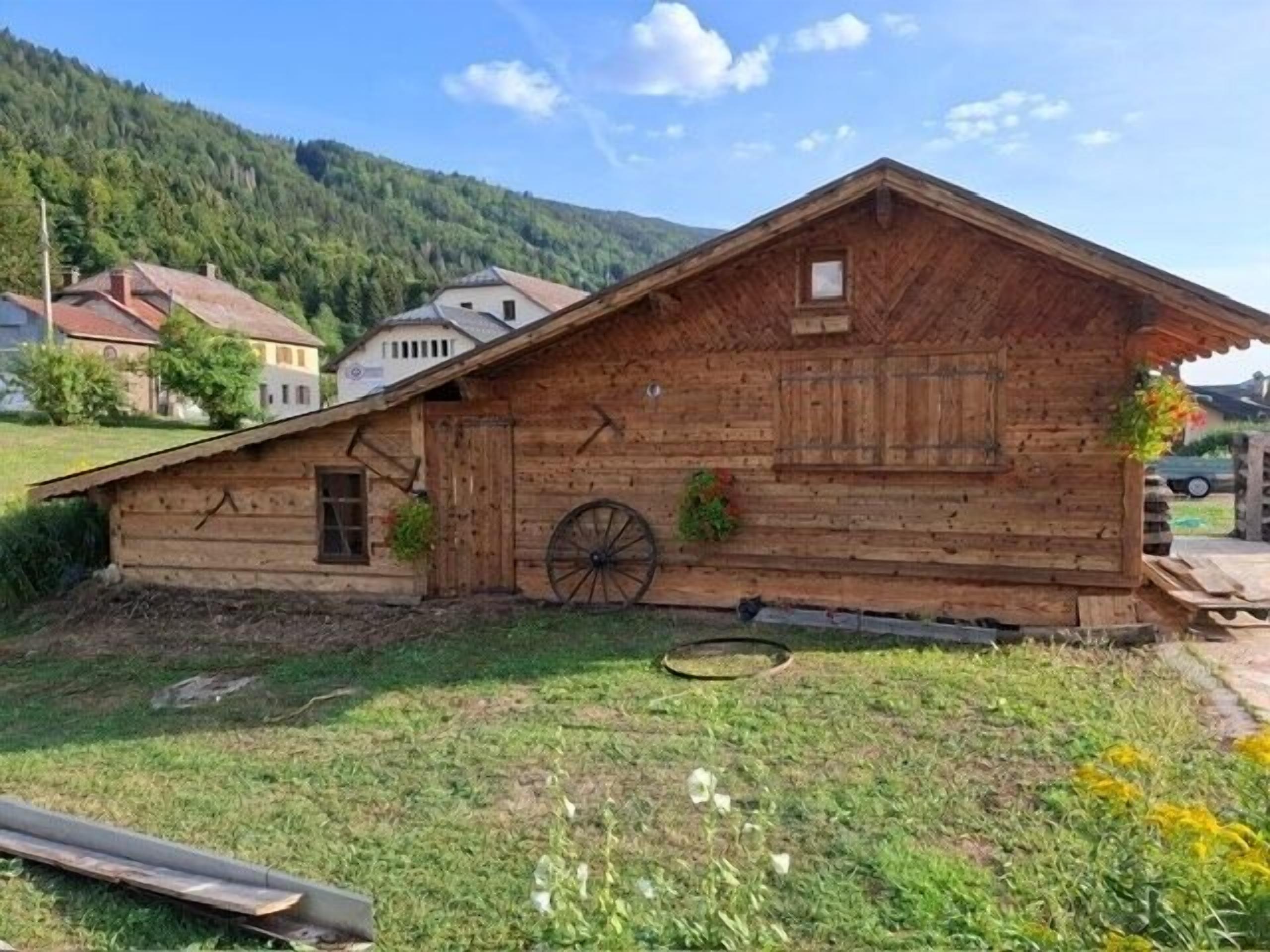 A wooden cabin with a wheel, surrounded by a grassy area and houses in the background.