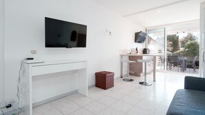 A modern room with a flat-screen TV, white furniture, a small trash bin, and a glass door leading to an outdoor area. - Le Nid d'Agde - Climatisé, à quelques pas du port (Agde)