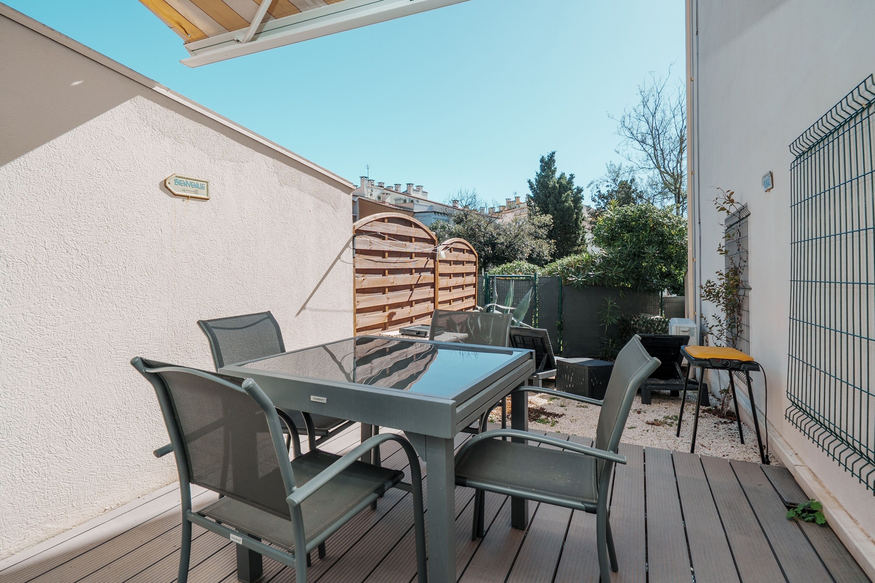 Een terras buiten met een glazen tafel en stoelen, een houten terras, een bakstenen muur en een metalen poort.