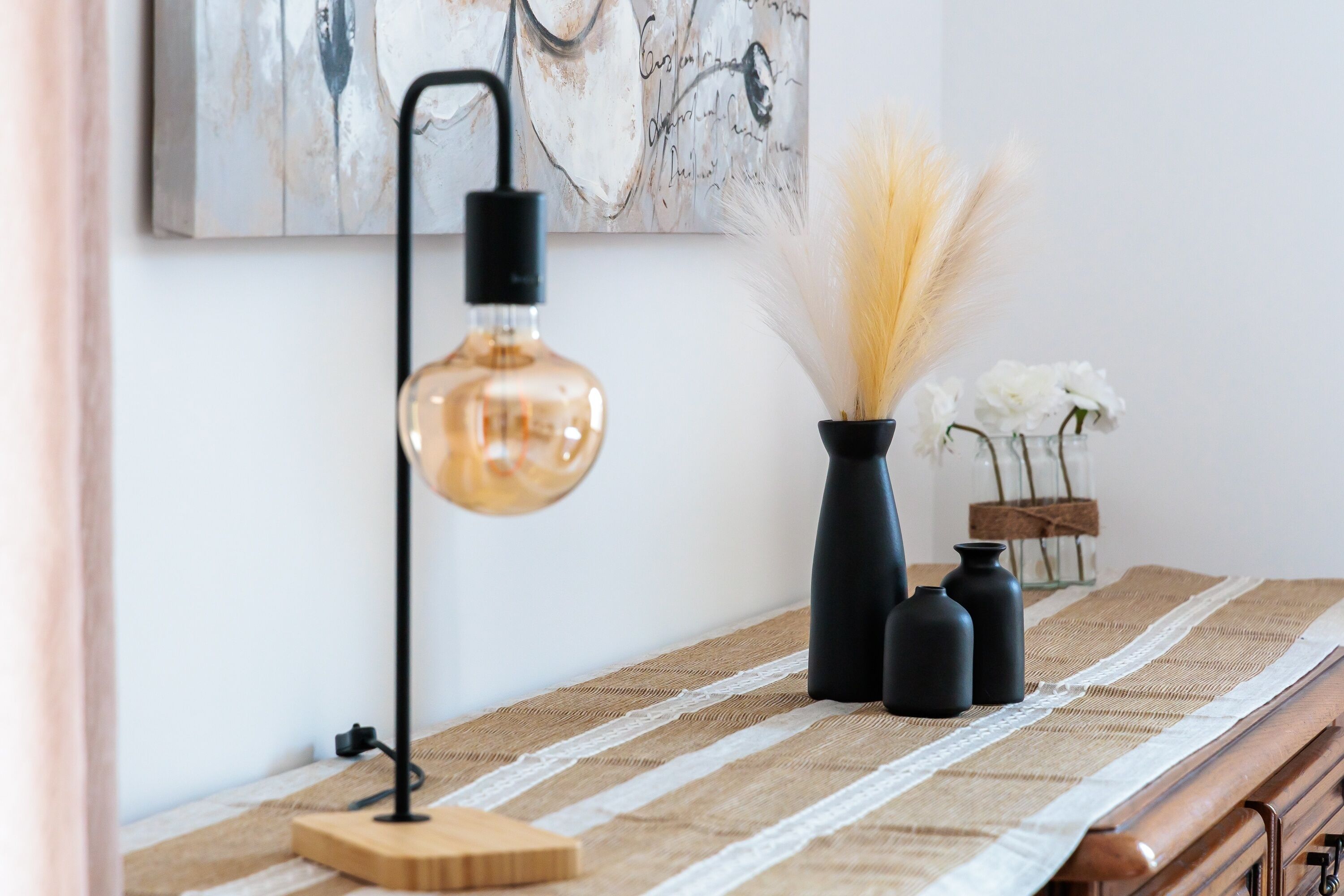 Une pièce comprenant une table, une lampe, un vase contenant des plantes séchées et une œuvre d’art encadrée accrochée au mur.