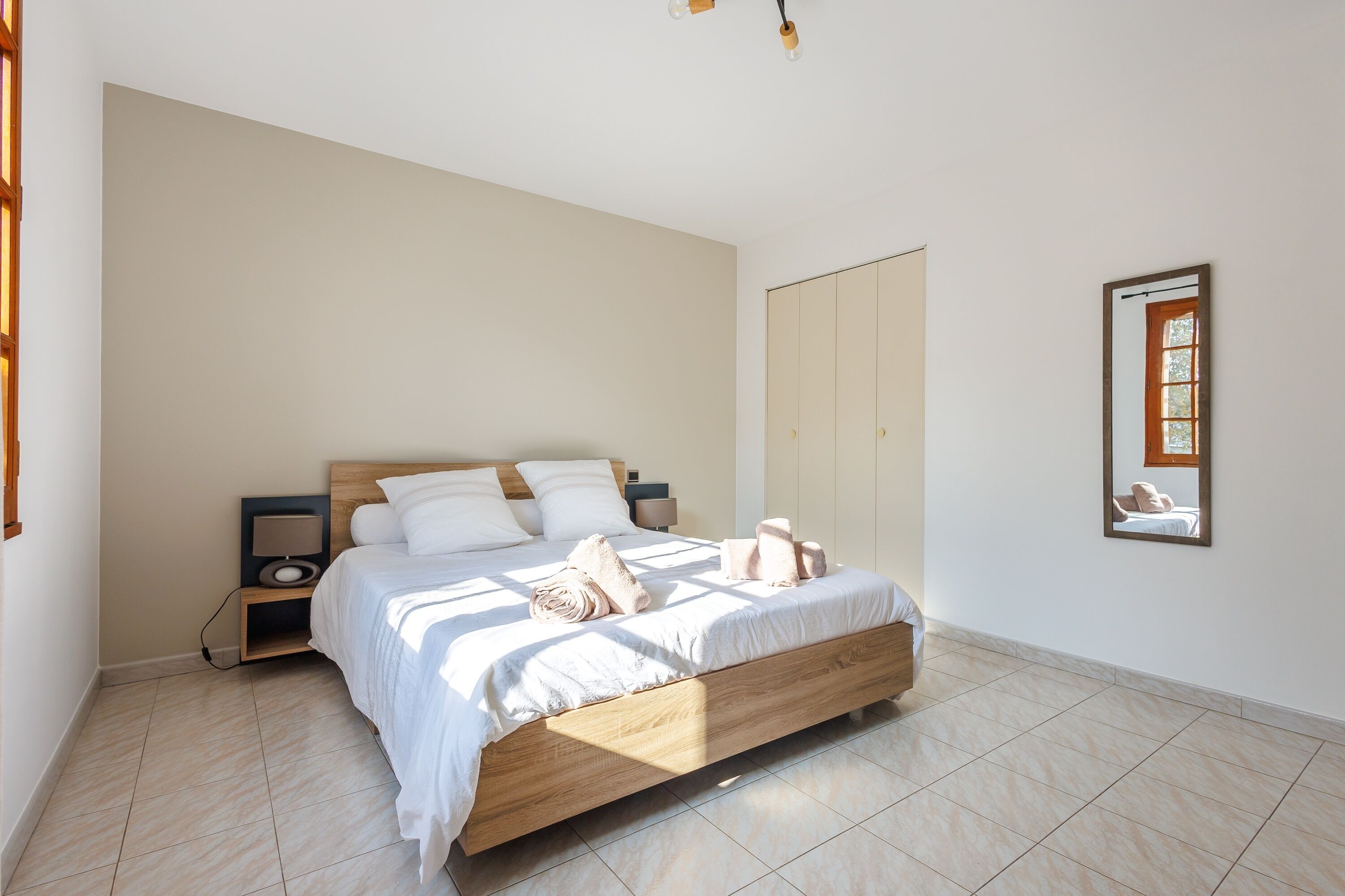 Een slaapkamer met een houten bed, witte beddengoed, een nachtkastje, een lamp en een spiegel aan de muur.