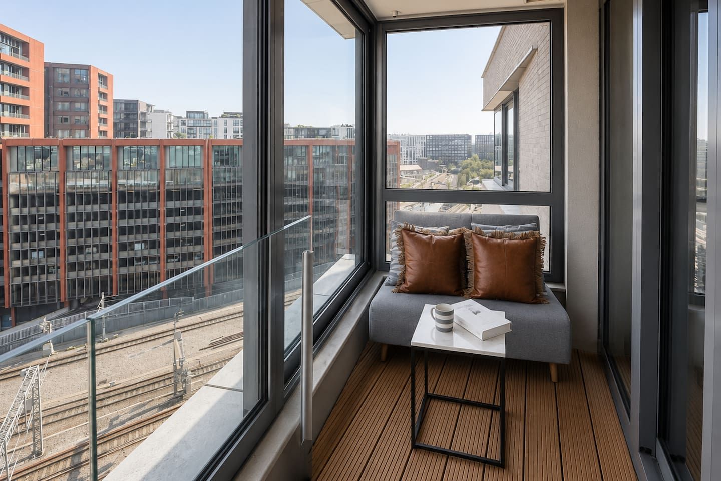 A modern balcony with a view of a cityscape, a cushioned seating area, and a small table with a book and a mug.