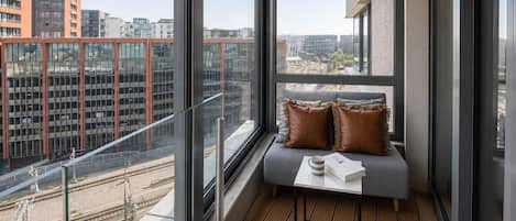 A modern balcony with a view of a cityscape, a cushioned seating area, and a small table with a book and a mug.