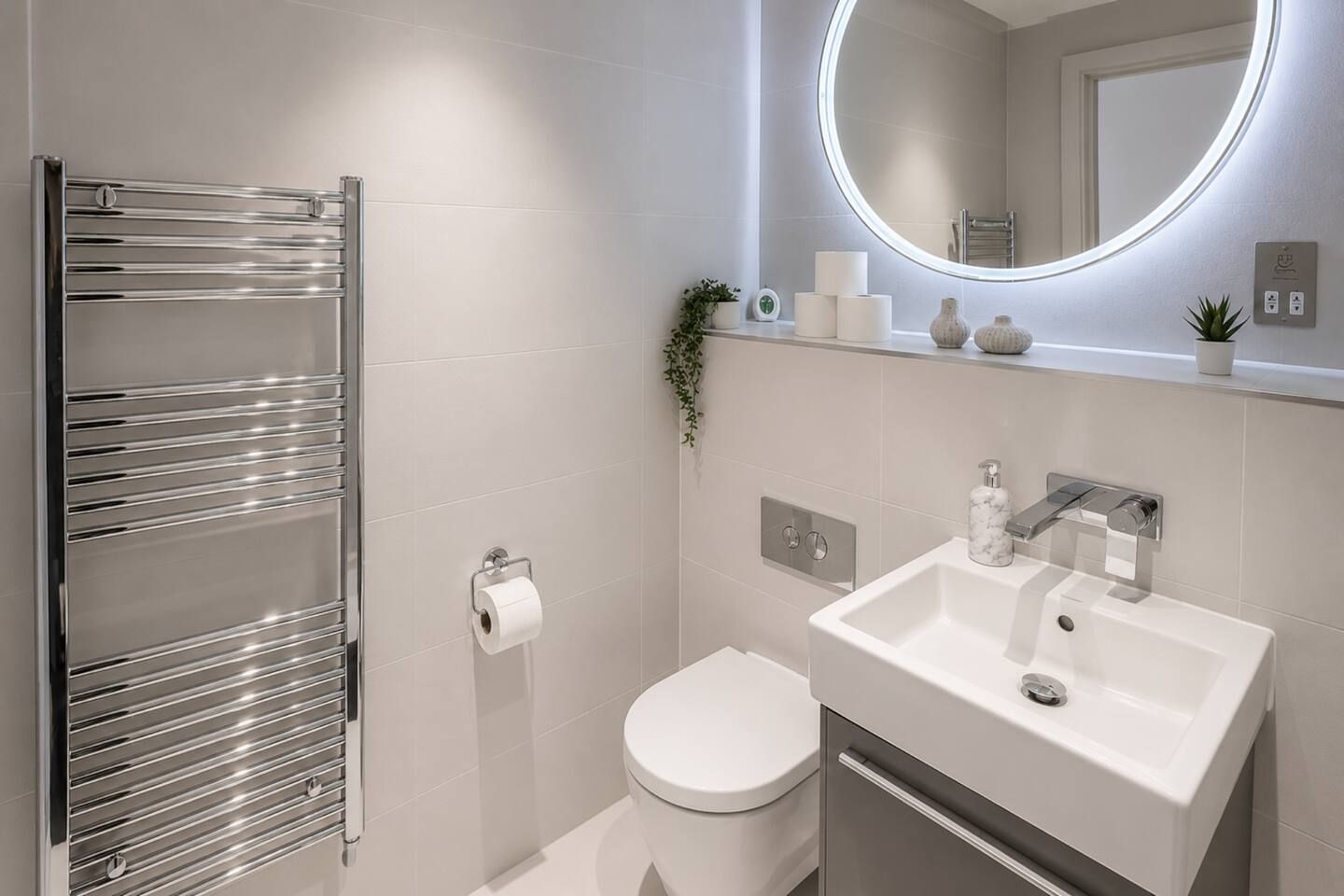Ein modernes Badezimmer mit einem chromfarbenen Handtuchhalter, einer Toilette, einem Waschbecken mit Seifenspender aus Marmor und einem Spiegel mit runder Beleuchtung.