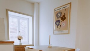 Un comedor con una mesa de madera, sillas blancas, un jarrón con flores y un cuadro colgado en la pared.