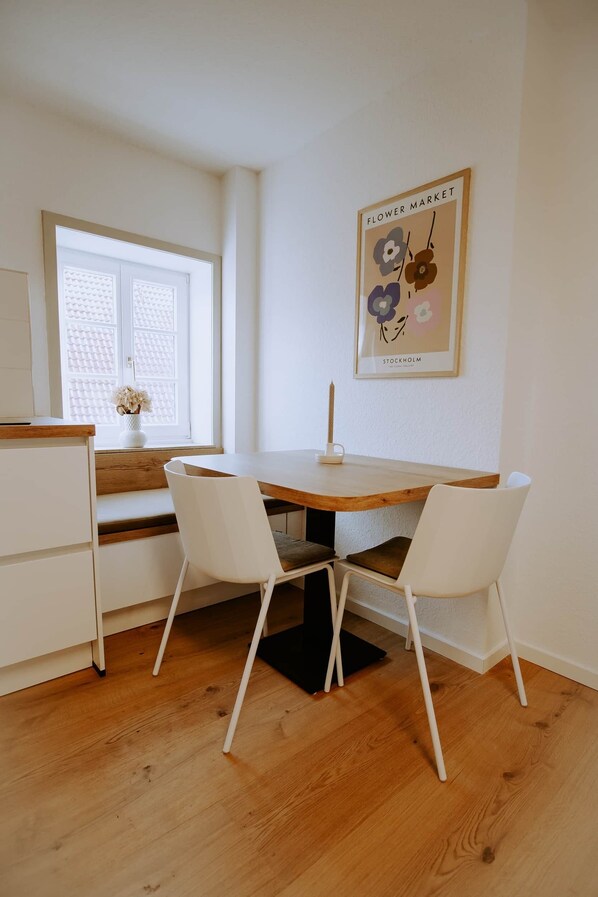 A dining area with a wooden table, white chairs, a vase with flowers, and a wall-mounted picture. - Leni's favorite place (County of Rietberg)