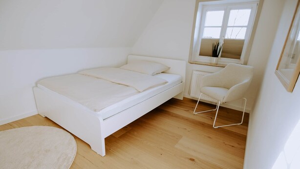 Una cama blanca en una habitación con suelo de madera, una silla blanca pequeña y una ventana con persianas.
