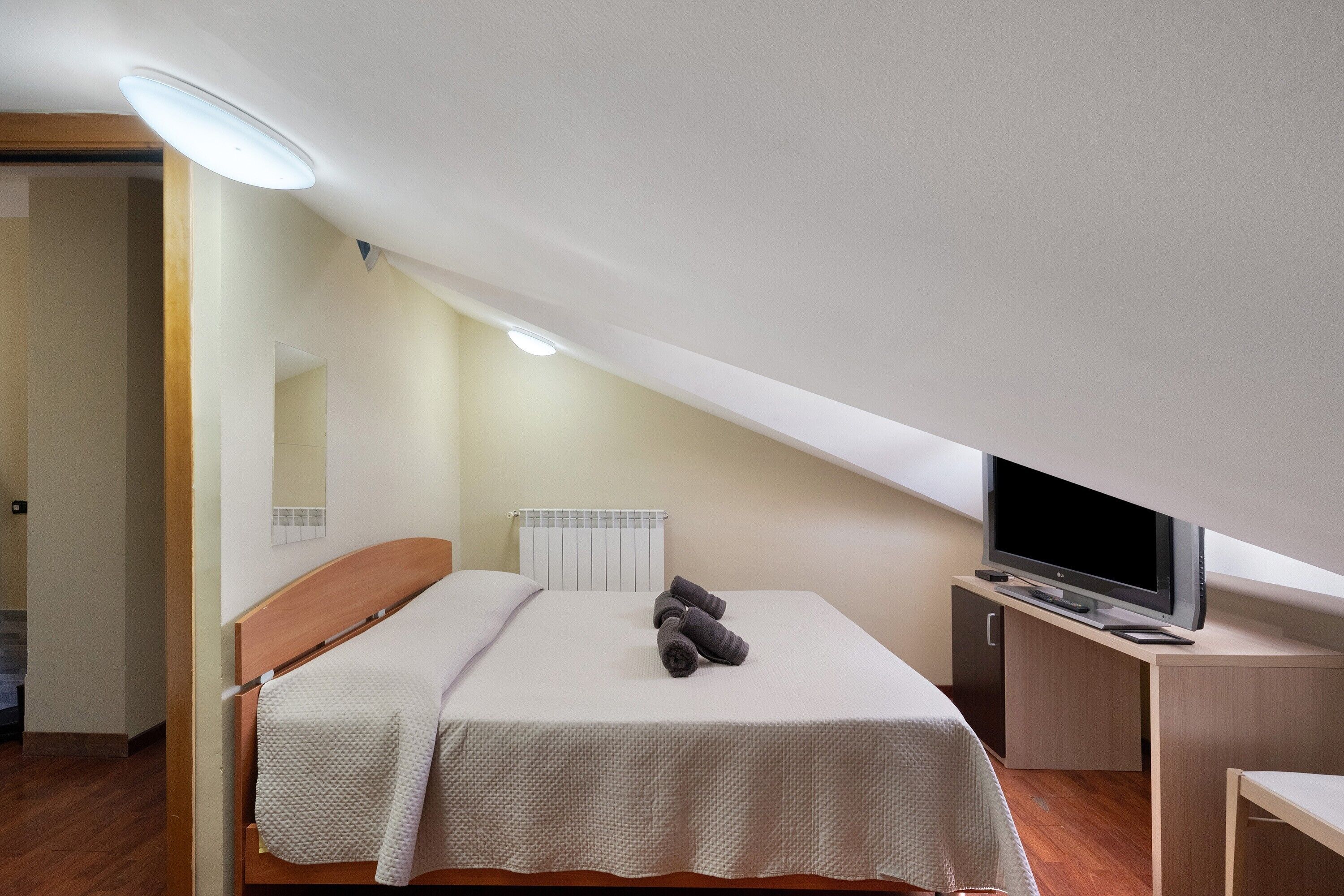 A bedroom with a bed, a TV on a stand, a radiator, and a door leading to another room.