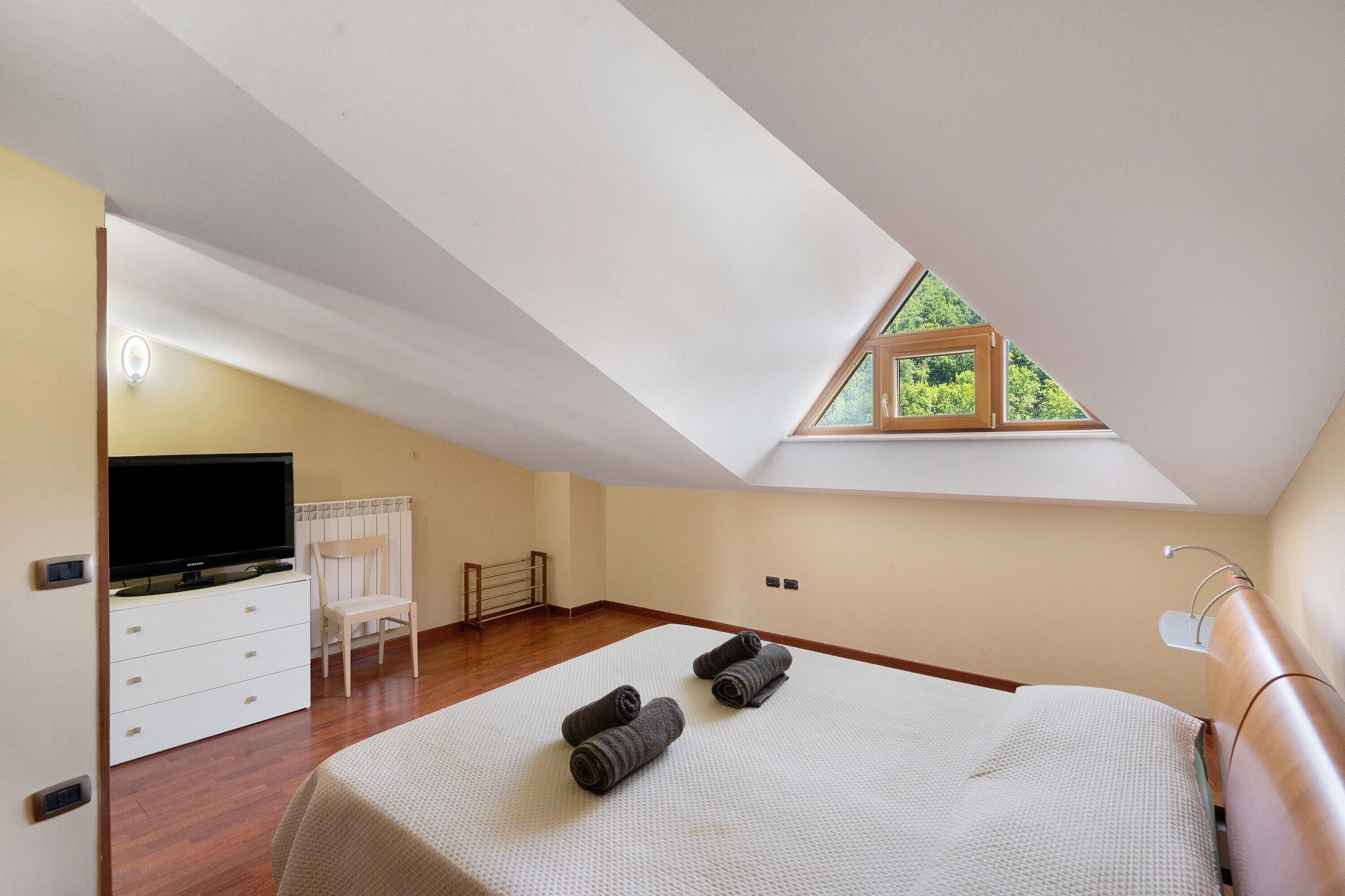 A bedroom with a sloped ceiling, a bed, a television, a chair, and a window with a view.