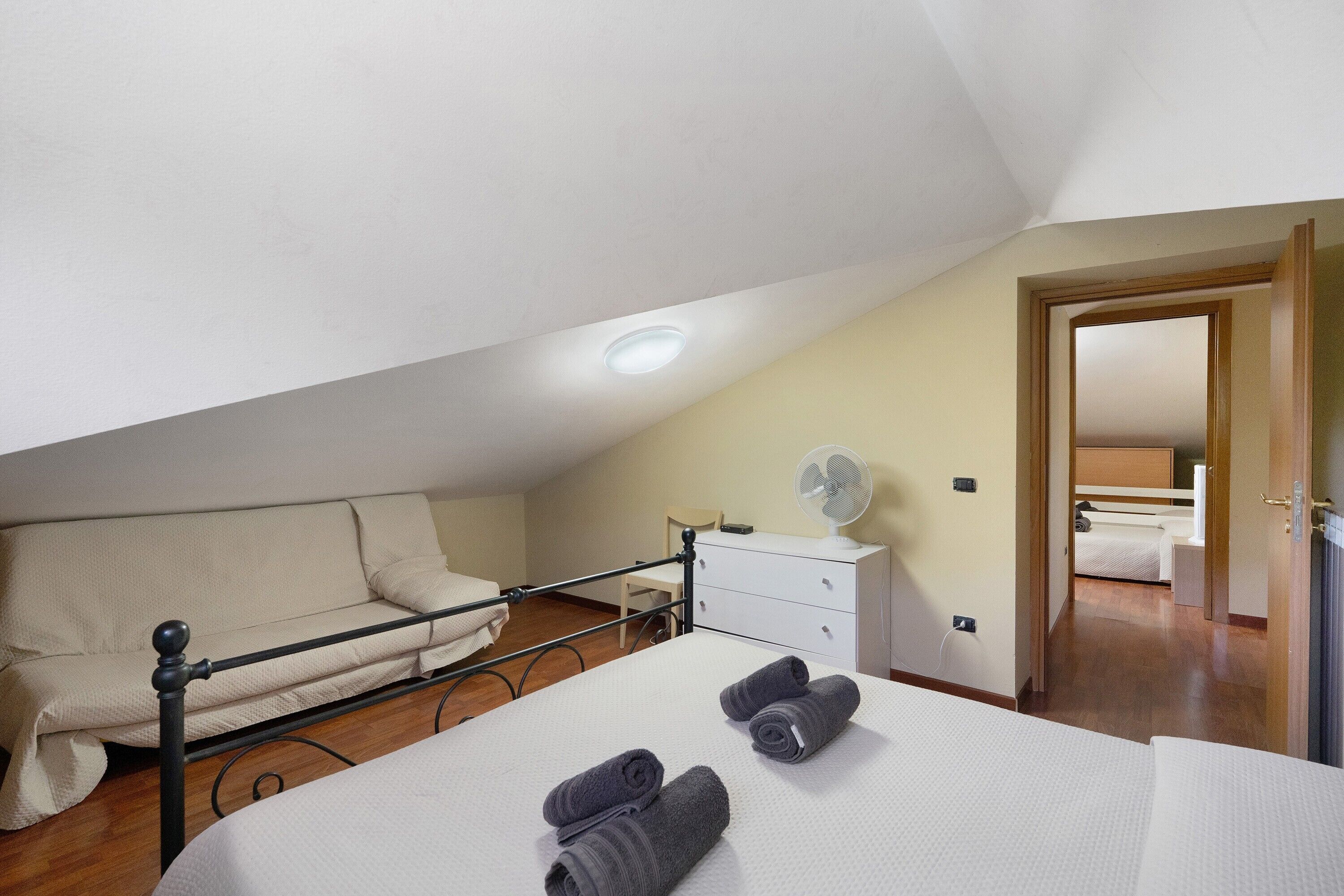A bedroom with a bed, a bedside table, a chair, a fan, and a door leading to another room.