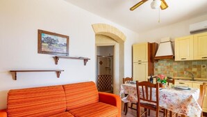 Una stanza con un divano, un tavolo da pranzo con sedie, un angolo cottura con pensili e una ventola a soffitto.