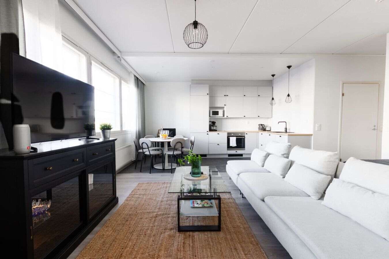 Een moderne woonkamer met een bank, salontafel en televisie. Er is een eethoek met stoelen en een tafel. De keuken is zichtbaar op de achtergrond, met kasten en apparatuur.